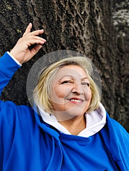 Confident beautiful mature woman stands with raised hands near a tree and smiles. Close up