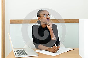Confident black business woman in glasses holding chin looking away at desk with notepad and laptop in office