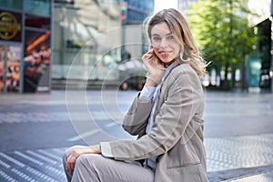 Confident blond businesswoman in suit, smiles at camera, sits outdoors near office buildings