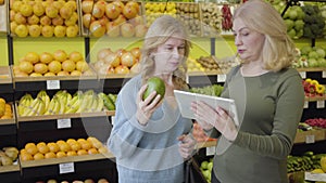 Confident blond Caucasian woman with tablet advising female friend choice of best fruit in grocery. Positive mature