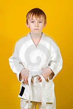 Confident boy in a white karate kimono over yellow background. Sports since childhood, discipline, first place, victory. Kids