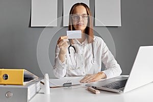 Confident brown haired woman doctor holding blank business or visit card nurse wearing white lab coat and stethoscope looking at