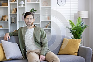 Confident brunette man in casual outfit leaning on back of sofa with bend arm in stylish interior. Handsome apartment