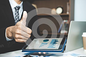 Confident business man in smart suit showing thumbs up and working on digital tablet and laptop at workplace desk