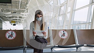 Confident business woman in a mask sitting at the airport, holding a passport and plane tickets, keeps a distance in a