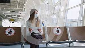 Confident business woman in a mask sitting at the airport, holding a passport and plane tickets, keeps a distance in a