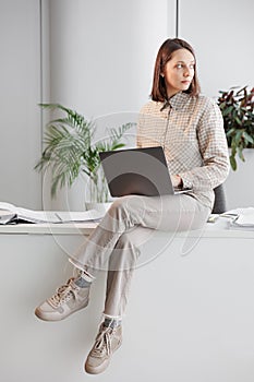 confident business woman is sitting on desk and working on laptop in office looking into distance