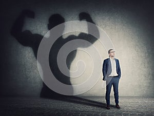 Confident businessman keeps hands in his pockets while casts a powerful person shadow on the wall behind. Business person