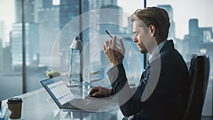 Confident Businessman in a Suit Sitting at a Desk in Modern Office, Using Laptop Computer, Next to