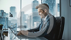 Confident Businessman in a Suit Sitting at a Desk in Modern Office, Using Laptop Computer, Next to