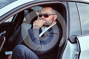 Confident businessman in sunglasses dressed in a formal suit sitting on the front seats in the luxury car.