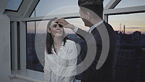 Confident businessman touching hair of beautiful brunette colleague standing on glazed balcony in the evening. Portrait