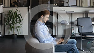 Confident businessman using laptop, sitting in armchair in modern office