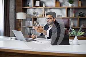 Confident businessman wearing suit and glasses sitting at desk with laptop during video chat