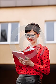 Confident businesswoman in glasses is doing notes in paper notepad by the office buildings background. Executive in red