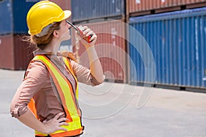 Confident of a businesswoman wearing a safety helmet and vest using radio communication in the container yard shipping area.