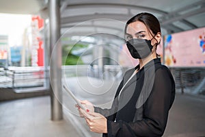 Confident businesswoman wearing suit and mask standing in city at walkway background. holding tablet outdoors and smile.