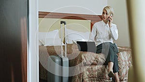 Confident businesswoman in white shirt using on laptop and talking at mobile phone gesturing while sitting on bed in