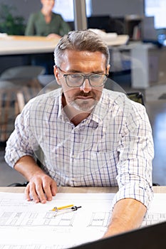 Confident caucasian male engineer using laptop while sitting with blueprint at desk in workplace