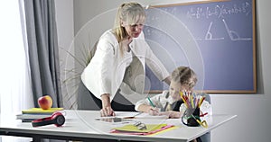 Confident caucasian middle aged tutor in formal suit standing next to schoolgirl and watching her writing in exercise