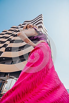 Confident Caucasian woman in trendy fashion poses on modern city building. Stylish outfit with shades of pink and purple