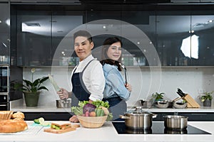 Confident chef and female commis posed to hold a knife with a soup ladle in cooking kitchen and standing back to back