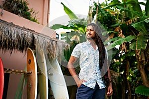 Confident dark-skinned surf instructor stands among colorful boards in tropical setting. BIPOC coach awaits students for