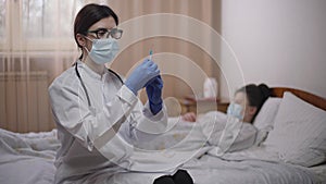 Confident doctor in coronavirus face mask with syringe posing in hospital ward on the left with blurred patient at