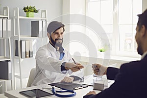 Confident doctor gives the patient a prescription while sitting at a table in a clinic office.