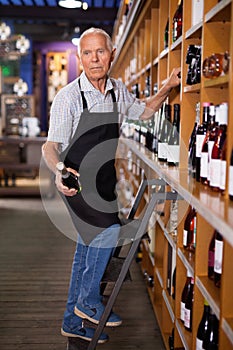 Confident elderly male owner of wine shop taking wine bottle from shelf rack and proffering to buy