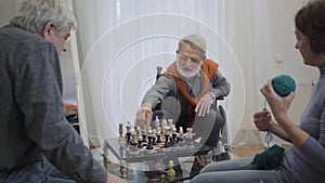 Confident elderly man in eyeglasses playing chess with fellow in nursing home as old woman knitting next to them