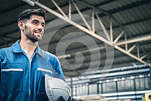 Confident engineer in blue jumpsuit holding hard hat looking forward in the warehouse.