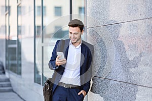 Confident enterpreneur checking messages on his mobile phone near office building wall in city