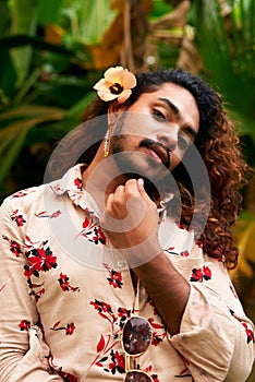 Confident ethnic man with flower in hair poses, tropical background. Gay BIPOC individual, curly hairstyle, floral shirt