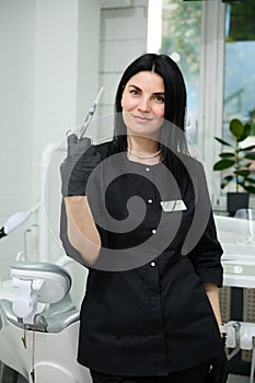 Confident female dental hygienist holding a syringe with dental anesthesia, looking at camera, standing by dentist chair