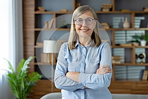 Confident female pensioner in pastel blouse standing with arms crossed on chest and smiling at camera. Mature lady in