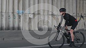 Confident fit focused cyclist riding a bicycle wearing helmet, black outfit and sunglasses. Bearded bike rider pedaling bicycle.