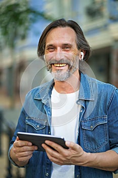 Confident freelancer man with grey haired hold digital tablet in hands having a call while standing on urban streets