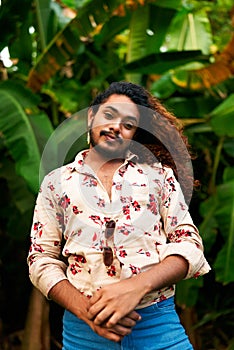 Confident gay man with curly hair stands in tropical setting. Flamboyant pose, bright floral shirt open, denim pants