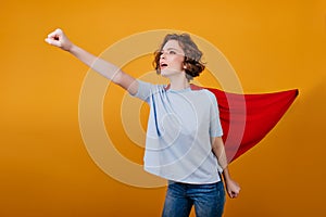 Confident girl in red mantle having fun during photoshoot in studio. Brave young woman in superhero attire waiting for
