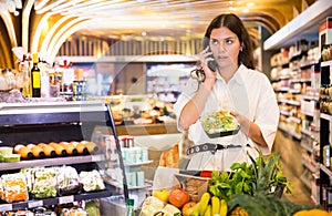 Confident girl in a supermarket is talking on a mobile phone, choosing frozen vegetables