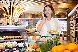 Confident girl in a supermarket is talking on a mobile phone, choosing frozen vegetables