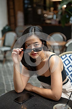 A confident and gorgeous Asian woman wearing sunglasses is sitting at a table in a cafe
