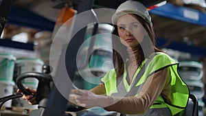 Confident gorgeous woman in hard hat putting hands on steering wheel sitting in forklift looking at camera. Portrait of