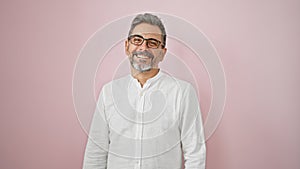 Confident, grey-haired hispanic man flaunting a charming smile, wearing glasses, standing over an isolated pink background,