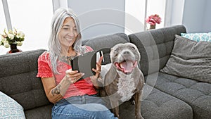 Confident grey-haired middle age woman blissfully using credit card on touchpad for online shopping, sitting with her dog on sofa