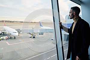 Confident handsome businessman on a business trip stands at the panoramic window in the waiting room of the international airport