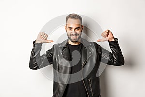 Confident handsome man wearing black leather jacket, pointing at himself and smiling self-assured, standing against