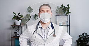 Confident happy Caucasian male surgeon doctor in lab coat, protective mask smiles at camera at modern hospital office.