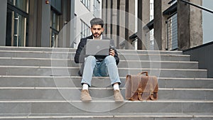 Confident hindu man in stylish clothes working on wireless laptop outdoors. Handsome young freelancer sitting on stairs